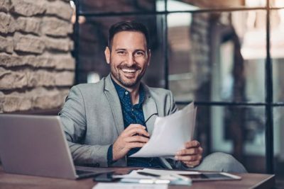 Wer ist in einem Unternehmen Verantwortlicher nach DSGVO? Hier symbolisiert durch einen jungen Mann, der mit Papieren in der Hand vor seinem, Laptop sitzt